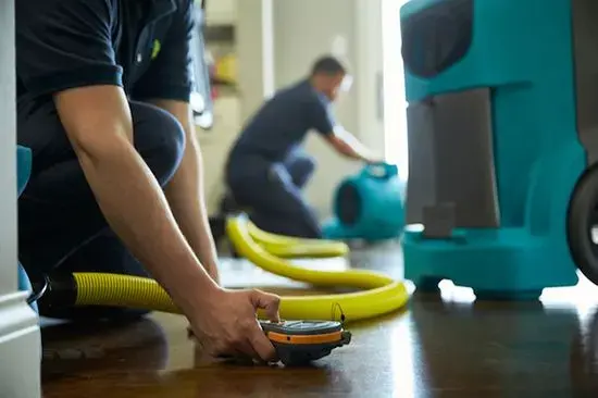 Technician kneeling with a moisture meter near a teal dehumidifier and yellow hose